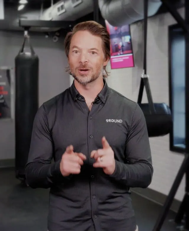 The owner of 9Round Franchising, the 24 hour gym franchise, stands with a punching bag in the background, wearing a dark shirt emblazoned with the 9Round logo, confidently pointing towards the camera.