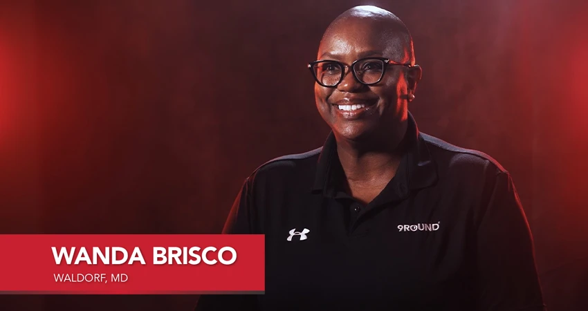 Person wearing glasses and a black polo shirt with 9ROUND logo, smiling against a dark background. Text on image: Wanda Brisco, Waldorf, MD. A glowing franchisee testimonial in every sense.