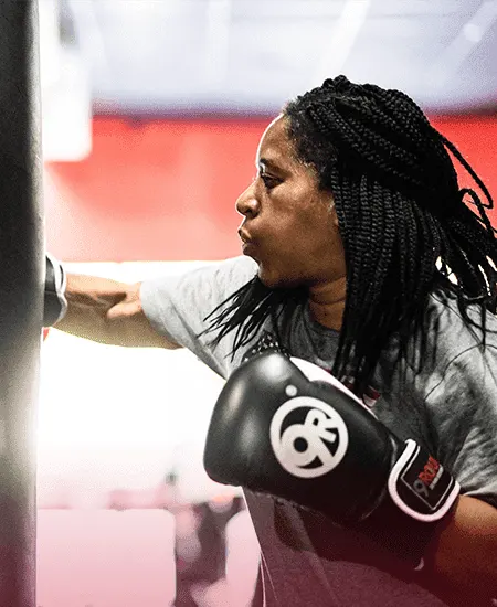 A person with braided hair, who loves fitness, delivers powerful punches to a punching bag in the gym while wearing boxing gloves.