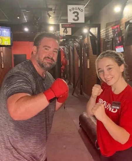 Two people wearing boxing gloves pose confidently in a bustling gym, standing proudly in front of a sign labeled Round 3.