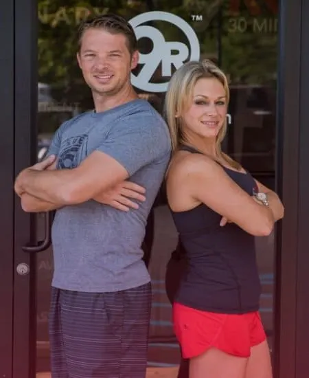 The owners of 9Round Franchising, LLC, stand back-to-back with arms crossed, smiling. Clad in casual athletic wear, theyre posed in front of a glass door featuring a gym logo, embodying the spirit of fitness franchising success.